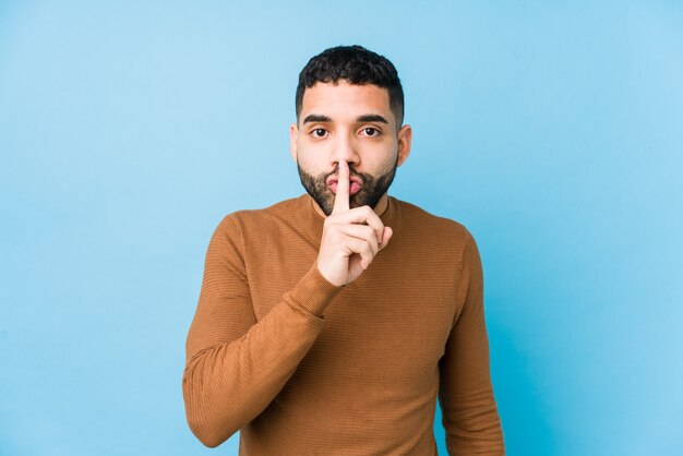 Joven latino contra una pared azul aislada manteniendo un secreto o pidiendo silencio.