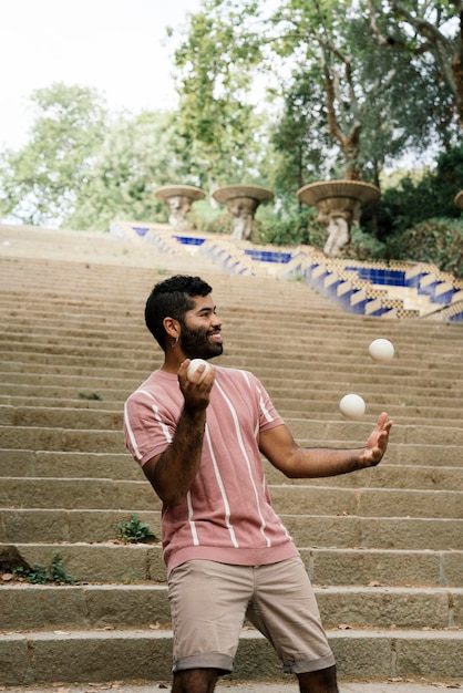 Foto joven latino haciendo malabares en las soleadas calles de barcelona haciendo un espectáculo con algunas bolas blancas mientras sonríe vestido con pantalones marrones y una camiseta de rayas pastel