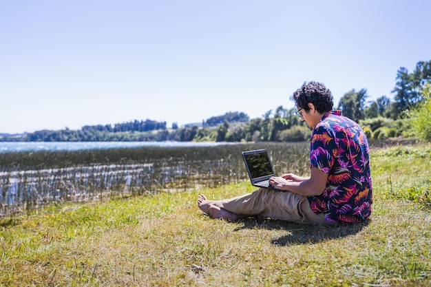 Joven latino haciendo trabajo remoto en línea desde el lago Concepto de teletrabajo Nómada digital