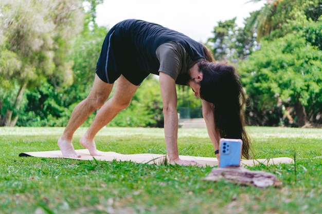 Joven latino hippie con cabello largo filmándose mientras se estira en una alfombra de yoga en un campo verde rodeado de árboles