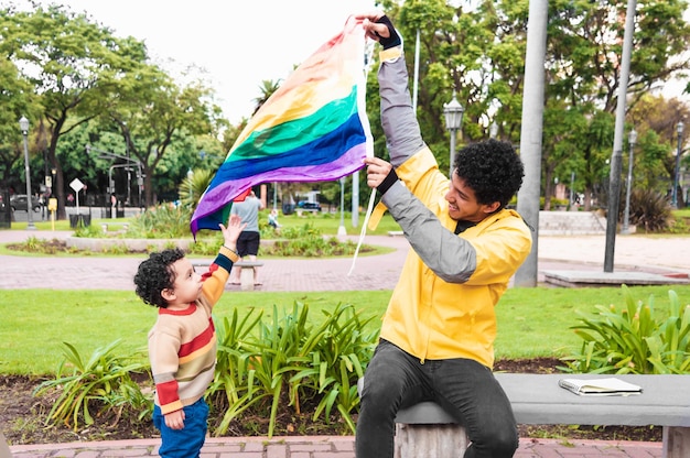 Joven latino negro, vestido de amarillo y con su hijo, en una plaza urbana, con una bandera gay, jugando y sonriendo, concepto lgbt