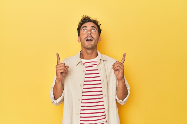 Joven latino posando sobre fondo amarillo apuntando al revés con la boca abierta