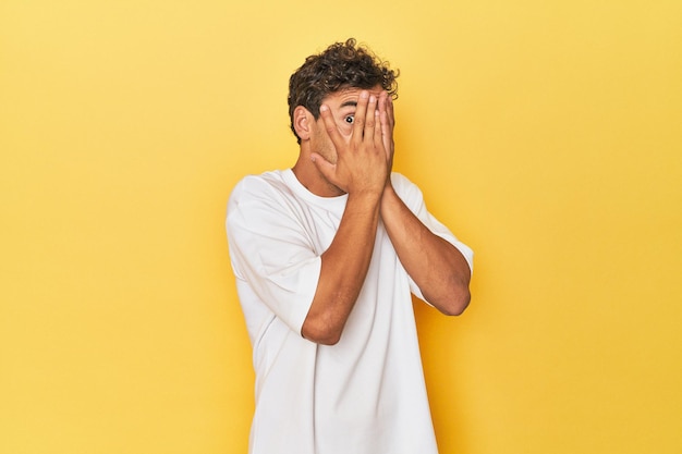 Foto joven latino posando sobre fondo amarillo parpadea entre los dedos asustado y nervioso
