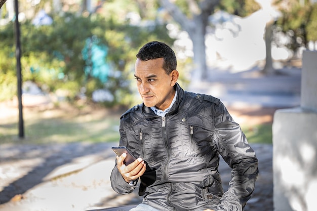 Joven latino sentado en un banco cuadrado usando su teléfono móvil