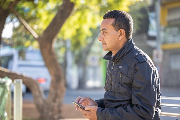 Joven latino sentado en un banco cuadrado usando su teléfono móvil