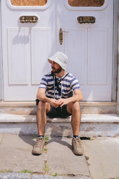 Joven latino con sombrero de cubo sentado frente a una puerta blanca en un barrio colonial Foto vertical