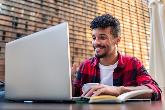 Joven latino sonríe haciendo una videollamada con su computadora portátil desde la terraza de una cafetería, el concepto de tecnología y estilo de vida urbano