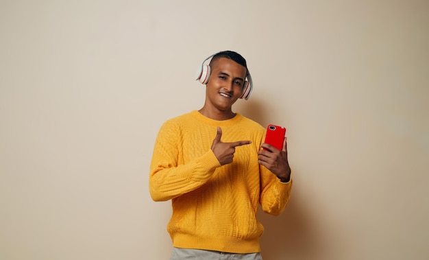 Un joven latino sonriendo con auriculares señalando con el dedo un teléfono móvil mirando a la cámara