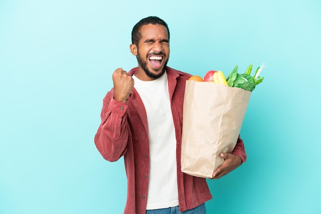 Joven latino sosteniendo una bolsa de compras aislado sobre fondo azul celebrando una victoria