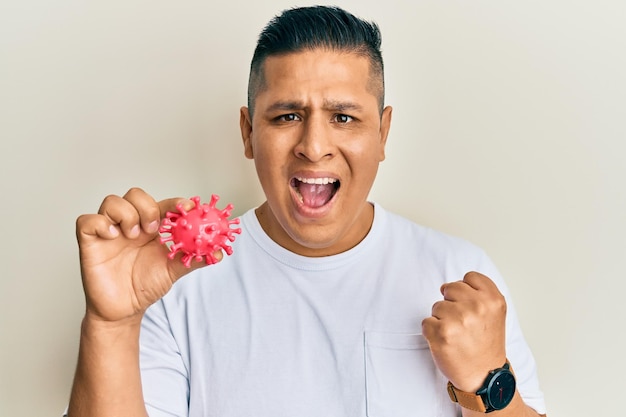 Joven latino sosteniendo un juguete de virus gritando orgulloso celebrando la victoria y el éxito muy emocionado con los brazos levantados