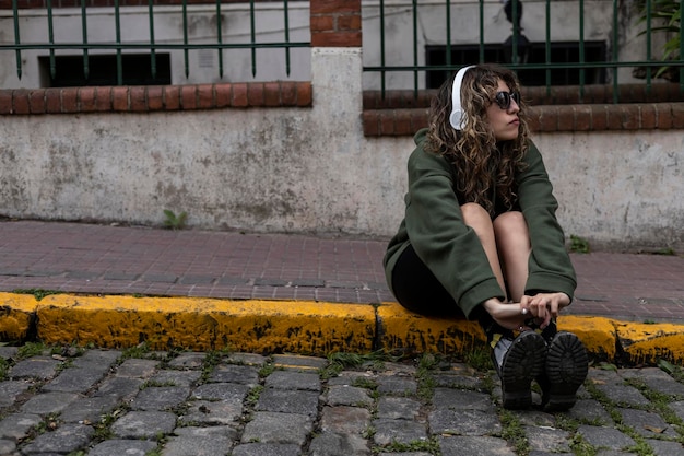 Joven latinoamericana de pelo rizado con gafas de sol sentada en la calle con los auriculares puestos