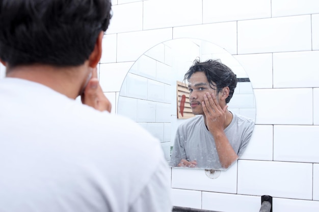 Joven lavándose la cara con jabón limpiador facial mirando el espejo en el baño en casa