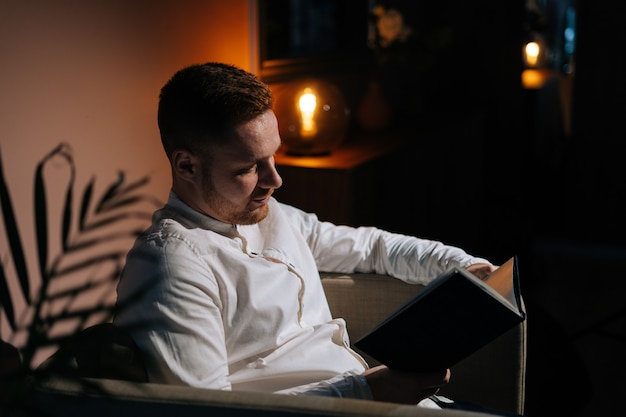 Foto joven lee el libro sentado en un sillón en la oscura sala de estar. concepto de relajación en casa.