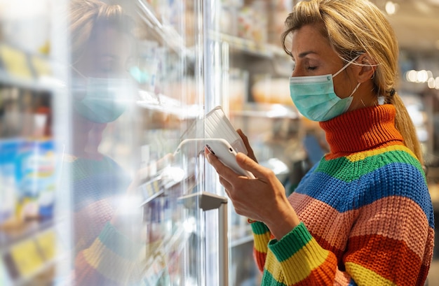 Joven lesbiana con suéter arcoiris con máscara facial usando teléfono móvil y comprando comestibles en la sección de congelados en el supermercado durante la pandemia de Coronavirus