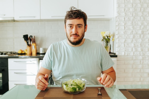 Un joven letárgico se sienta en la cocina frente a un plato de verduras con las manos sobre la mesa. Un joven con camiseta blanca mira a la cámara con decepción por la dieta Disparando en casa