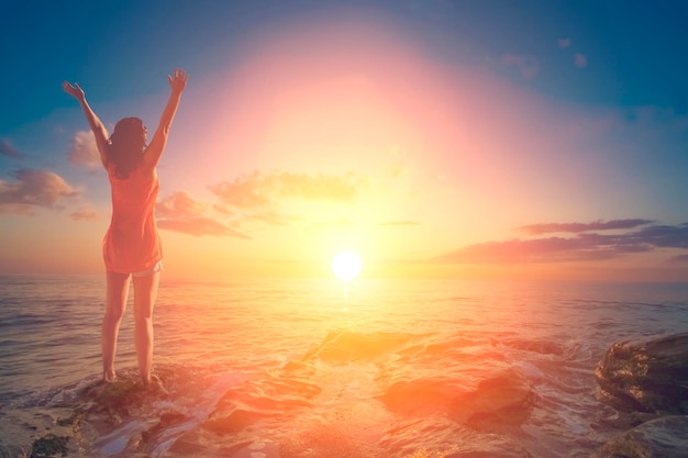 La joven levantó la mano hacia el sol contra las rocas del mar y el cielo con nubes.