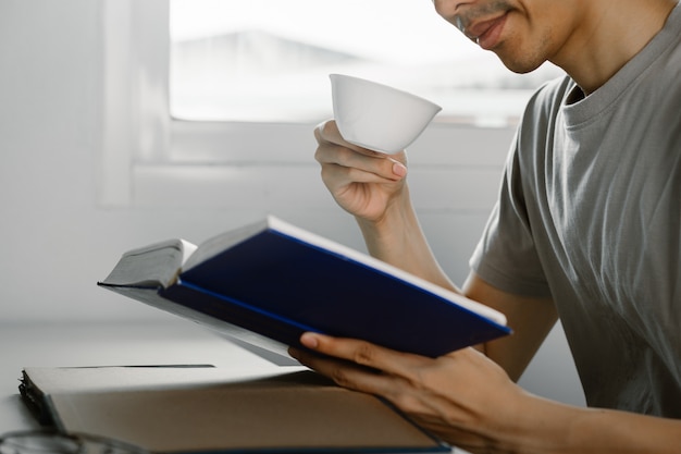 Joven leyendo un libro y bebiendo té en el escritorio en el tiempo libre de trabajar en casa, el conocimiento y el concepto de aprendizaje