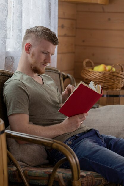 Joven libro de lectura con tapa roja sobre banco de mimbre en casa de madera de campo