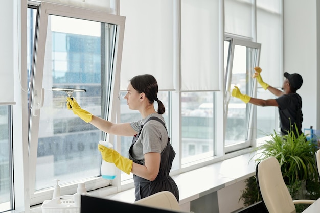 Joven limpiadora en ropa de trabajo lavando grandes ventanas de oficina de espacio abierto