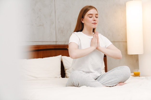 joven linda chica complacida en el interior de su casa en el dormitorio meditar en la cama.