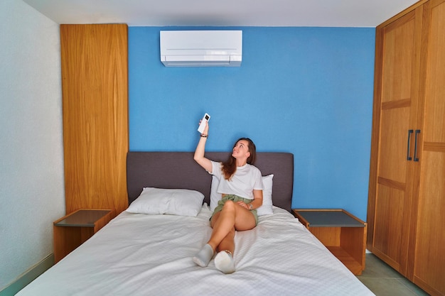 Joven linda chica sonriente acostada en la cama y ajustando la temperatura del aire acondicionado usando el control remoto en el dormitorio