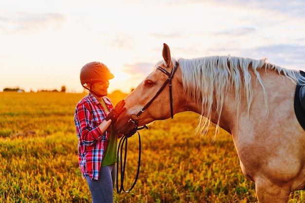 Joven linda feliz alegre satisfecha mujer sonriente jinete con casco abrazando y acariciando hermoso caballo palomino rubio en el prado al atardecer a la hora dorada