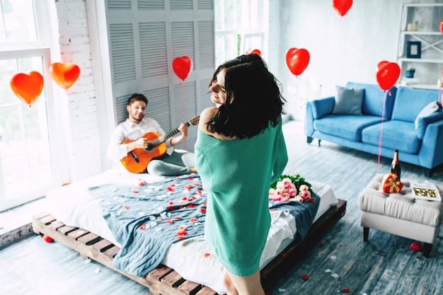 Una joven linda y hermosa mujer está bailando en medio del dormitorio mientras su chico guapo toca la guitarra para ella. Amor, celebración, romántico, st. Día de San Valentín, día de la mujer, cumpleaños, fecha.