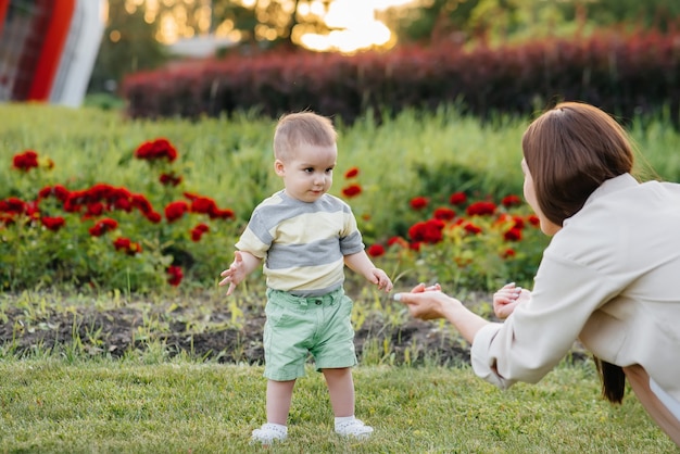 Una joven linda madre ayuda y enseña a su pequeño hijo a dar sus primeros pasos durante la puesta de sol en el parque sobre el césped.