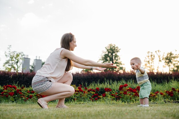 Una joven linda madre ayuda y enseña a su pequeño hijo a dar sus primeros pasos durante la puesta de sol en el parque sobre el césped.