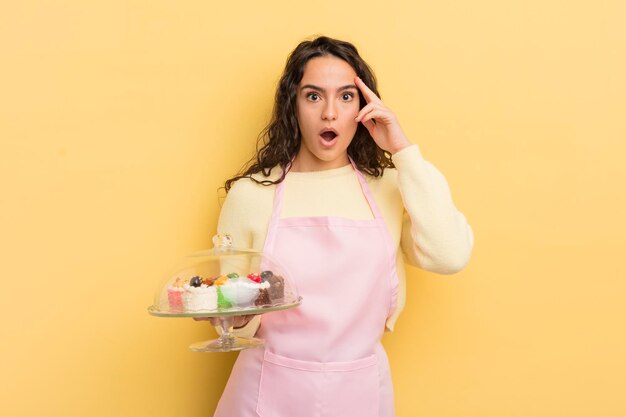 Joven linda mujer hispana que parece sorprendida al darse cuenta de una nueva idea de pensamiento o concepto de concepto de chef y pasteles