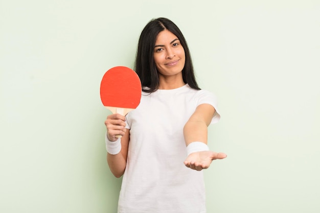 Joven linda mujer hispana sonriendo felizmente con amabilidad y ofreciendo y mostrando un concepto de ping pong