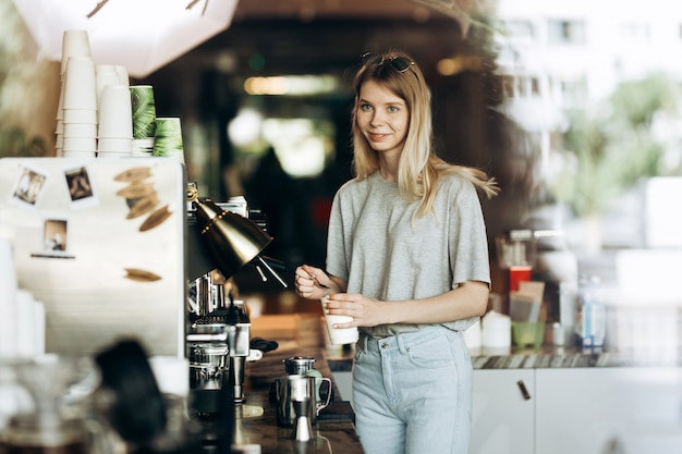 Una joven linda rubia delgada con cabello largo, vestida con ropa informal, está cocinando café en una cafetería moderna. Se muestra el proceso de elaboración del café. .