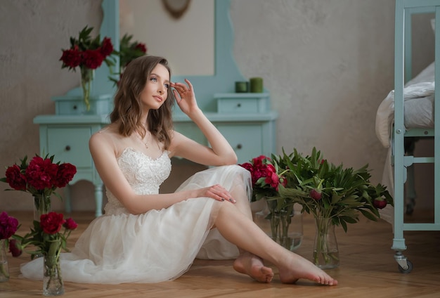 Una joven linda con un vestido blanco hasta la rodilla sentada en el suelo en una habitación con flores.