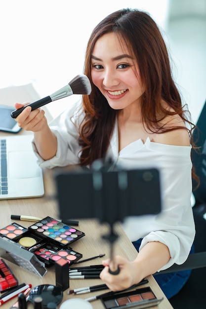 Joven y linda vloguera, influenciadora o vendedora en línea asiática que sostiene un teléfono inteligente en un palo largo para tomarse un video selfie y transmitir una transmisión en vivo para una revisión cosmética. Concepto de marketing online.