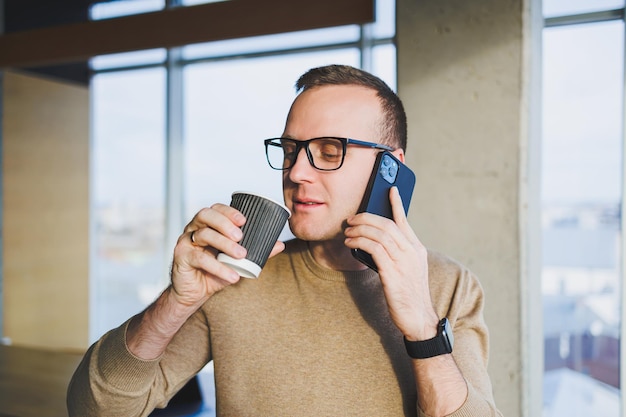 Un joven lindo con un suéter marrón y anteojos está hablando emocionalmente por un teléfono móvil y bebiendo café mientras se relaja en la oficina Un joven autónomo trabaja de forma remota