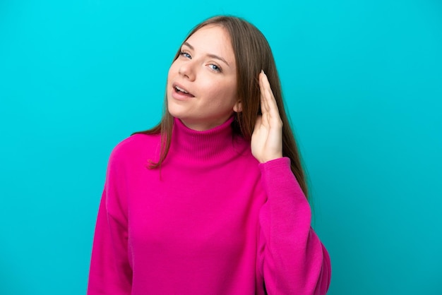 Foto joven lituana aislada de fondo azul escuchando algo poniendo la mano en la oreja