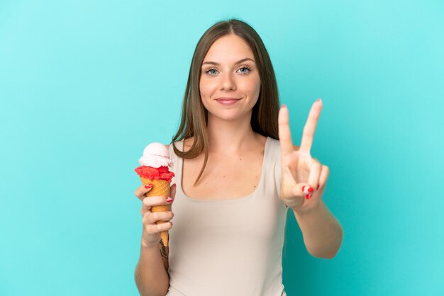 Joven lituana con helado de cucurucho aislado de fondo azul sonriendo y mostrando el signo de la victoria