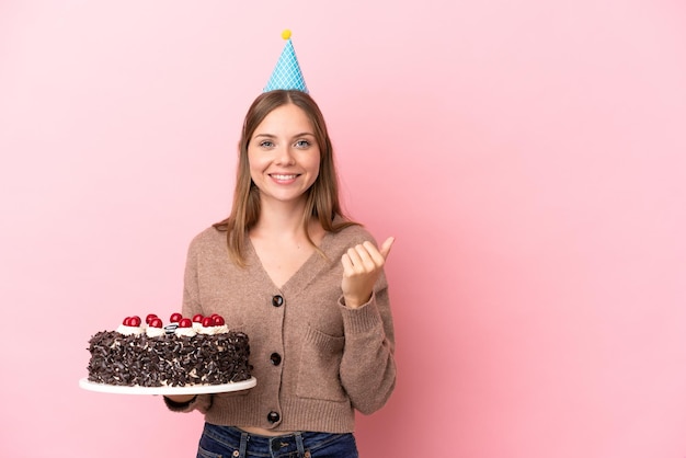 Joven lituana sosteniendo un pastel de cumpleaños aislado en un fondo rosa apuntando hacia un lado para presentar un producto