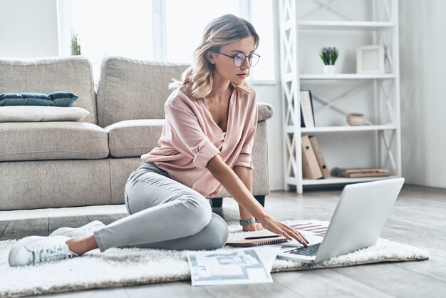 Joven y lleno de ideas. Mujer joven pensativa en gafas trabajando usando la computadora mientras el piso en casa