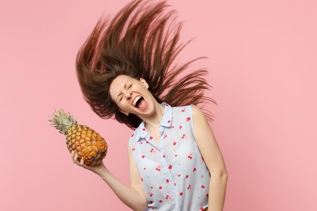 Una joven loca y gritadora con el pelo suelto manteniendo los ojos cerrados sostiene fruta de piña madura fresca aislada en un fondo rosa pastel. El estilo de vida vívido de la gente relaja el concepto de vacaciones. Simulacros de espacio de copia.