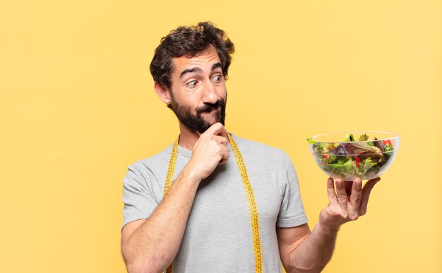 Joven loco barbudo haciendo dieta expresión de pensamiento y sosteniendo una ensalada