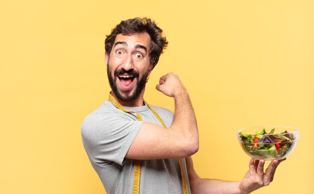 Joven loco barbudo haciendo dieta expresión de sorpresa y sosteniendo una ensalada