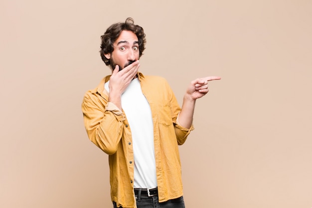 Joven loco loco, sintiéndose feliz, sorprendido y sorprendido, cubriéndose la boca con la mano y apuntando al espacio de copia lateral en la pared plana