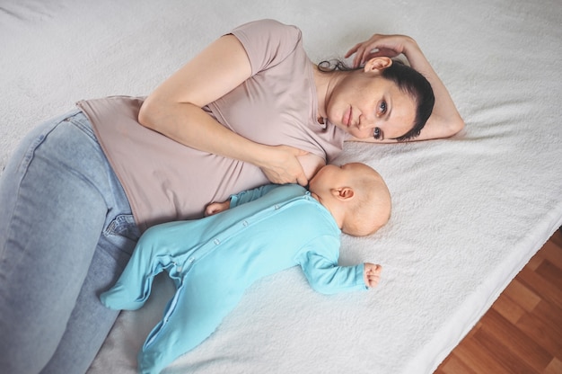 Joven madre se acuesta con un bebé recién nacido lindo bebé en mono azul en la cama, lo abraza y lo amamanta con leche materna