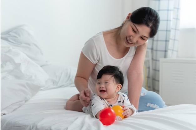 Joven madre asiática hermosa con bebé asiático en la cama y jugando a la pelota de juguete juntos en la cama blanca con sentirse feliz y alegre y el bebé que se arrastra en la cama. Concepto de familia bebé