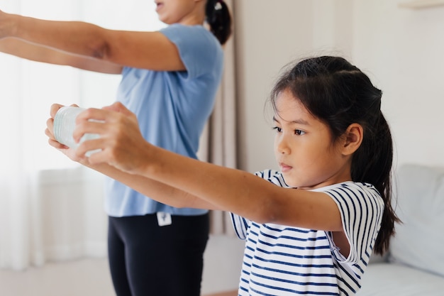 Joven madre asiática y su hija haciendo ejercicios de yoga de estiramiento y usando la botella de agua para hacer mancuernas juntos en casa en la rutina diaria.