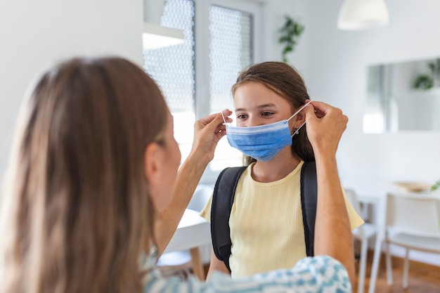 La joven madre ayuda a su hija con una máscara médica a prepararse para ir a la escuela. Evitar el brote de Covid-19 o coronavirus. De vuelta a la escuela