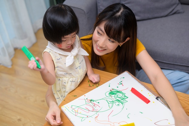 Una joven madre ayudando a su hija a dibujar con lápices de colores en la sala de estar de casa.