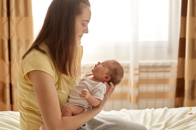 Joven madre de cabello marrón sosteniendo a su pequeño bebé mientras estaba sentada en su acogedora habitación en la cama abrazando a su lindo bebé recién nacido en casa expresando amor y ternura