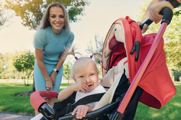 Joven madre caminando en el parque con bebé sonriente en cochecito de verano rojo. Manos del padre sosteniendo el cochecito.